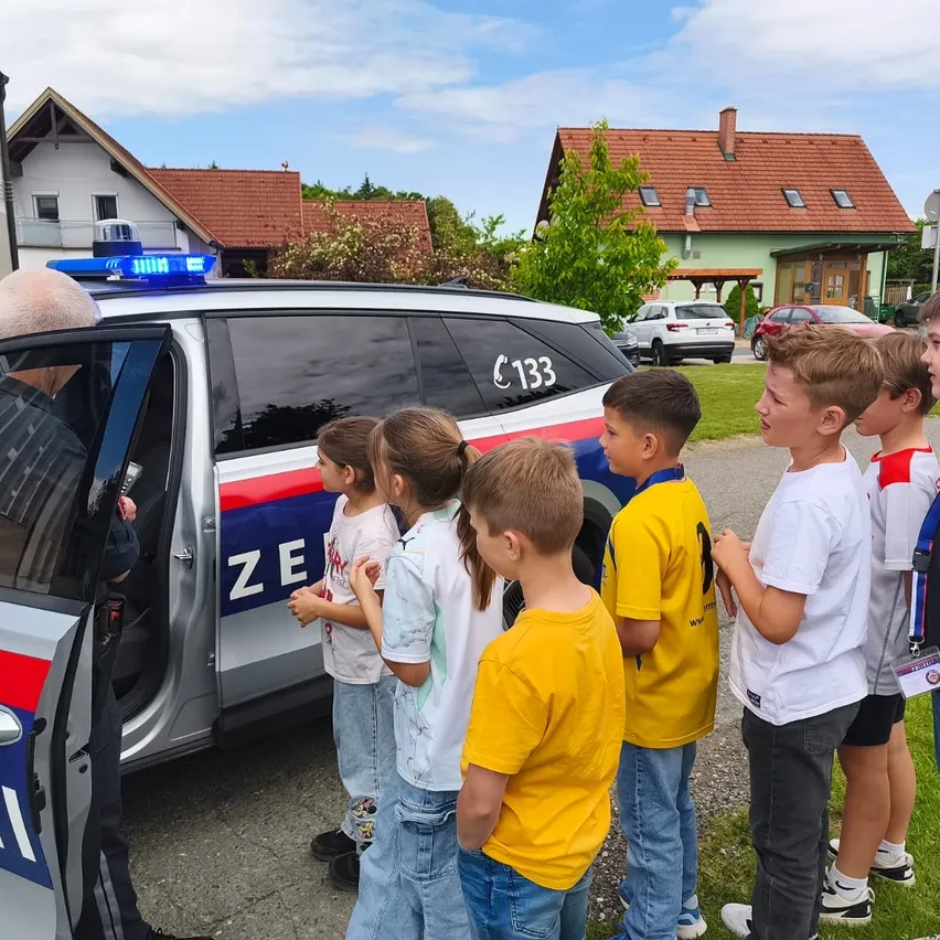 Eine Gruppe von Kindern steht um einen Polizeiwagen. Das Auto hat die Nummer 133. Ein Polizist spricht mit ihnen. Hinter dem Auto stehen Häuser und geparkte Autos.