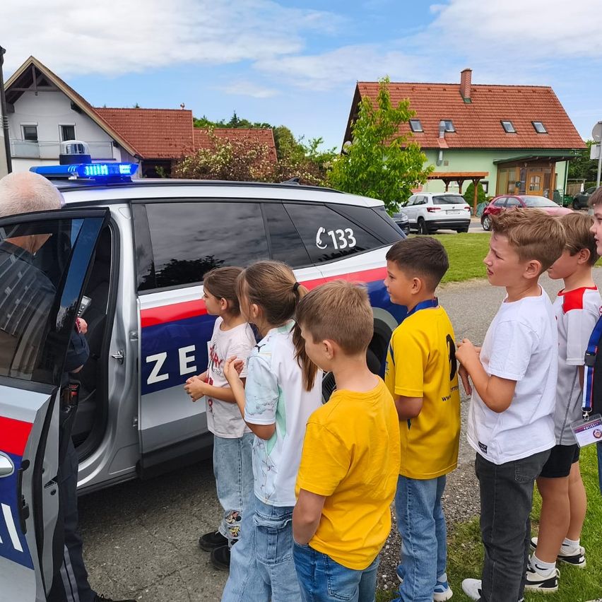 A group of children stands around a police car. The car has the number 133. A police officer talks to them. Behind the car, there are houses and parked cars.