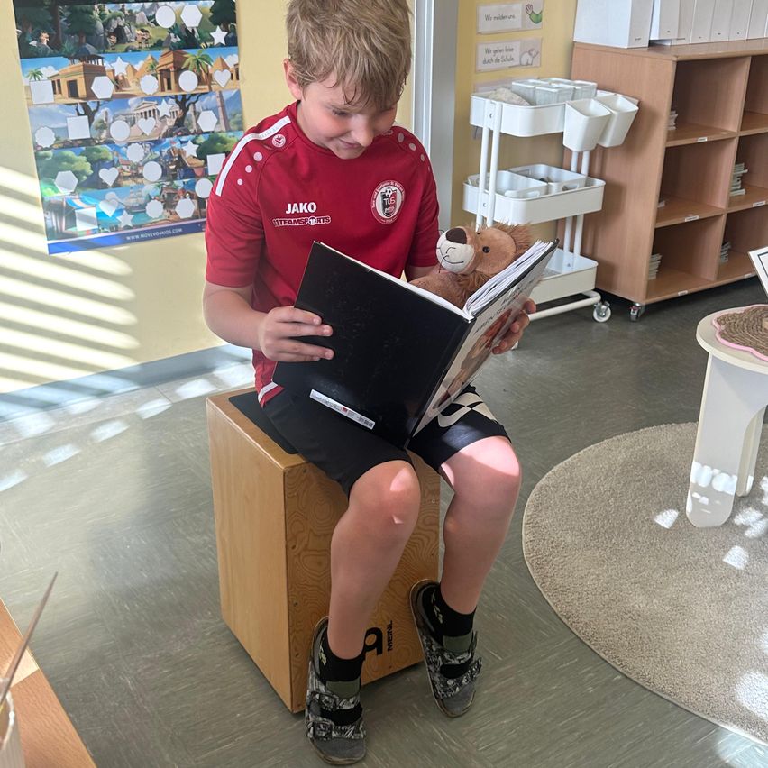 Ein junger Junge in einem roten Shirt sitzt auf einem Holzhocker und liest ein Buch, während er einen Teddybär hält. Er befindet sich in einem Raum mit einem Bücherregal und einem Teppich.