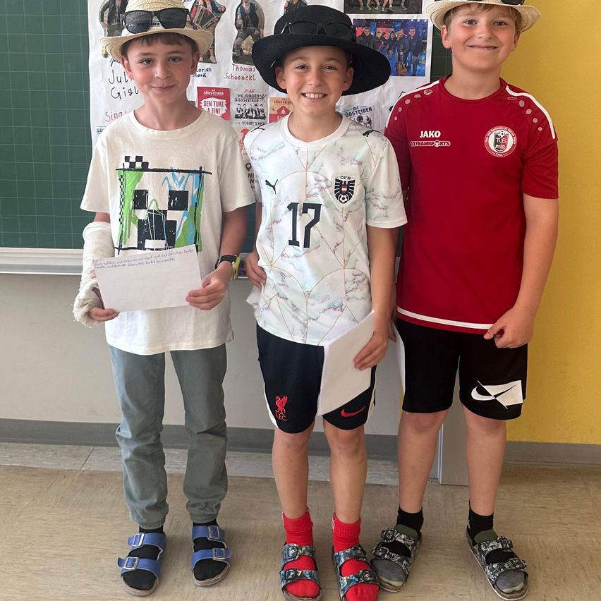 Three boys stand in a classroom, smiling. The boy on the left holds a paper. The middle boy wears a jersey with the number 17. The boy on the right wears a red jersey with a logo. They wear hats and sandals.