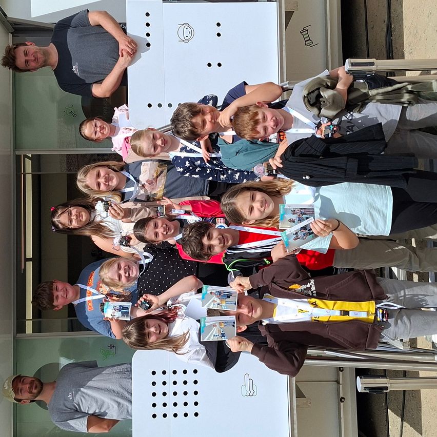 A group of children and adults pose for a photo outside a building with a smiling face. They hold various items like badges and booklets.