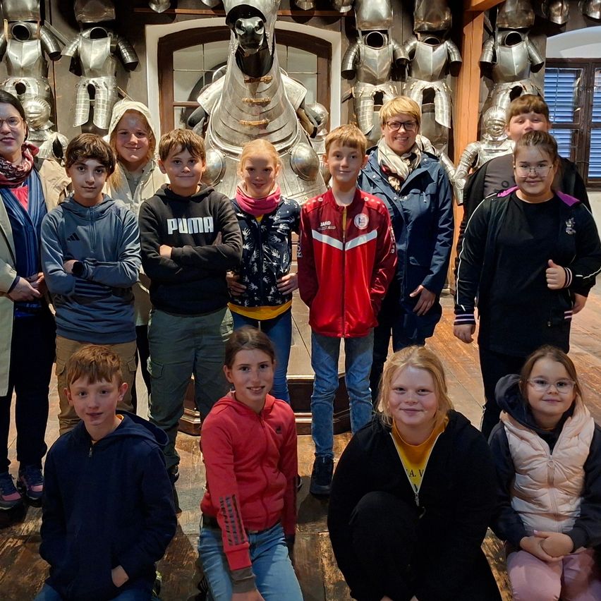 A group of children and adults posing in front of a wall with various knight armor on display. The children are smiling, some wearing glasses. The adults are standing behind the children.