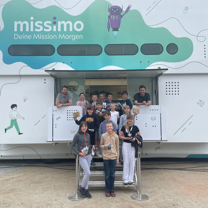 Kinder und Erwachsene stehen vor einem mobilen Lernfahrzeug. Einige halten Bücher und Auszeichnungen. Das Fahrzeug hat ein buntes Design mit dem Text 'missimo' und 'Deine Mission Morgen.'