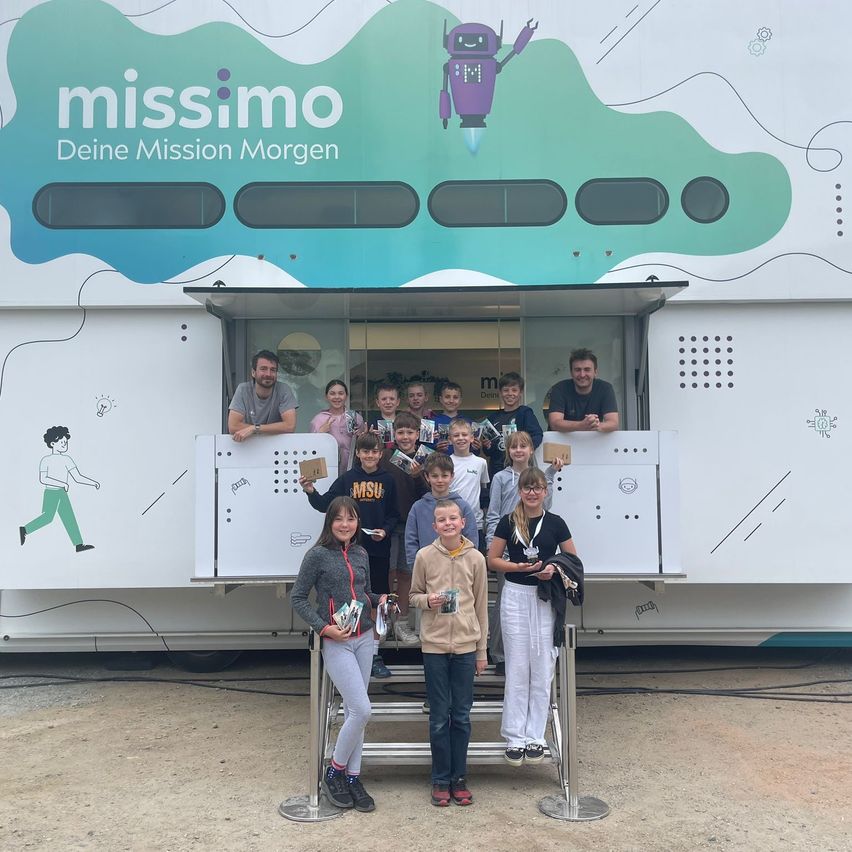 Kinder und Erwachsene stehen vor einem mobilen Lernfahrzeug. Einige halten Bücher und Auszeichnungen. Das Fahrzeug hat ein buntes Design mit dem Text 'missimo' und 'Deine Mission Morgen.'