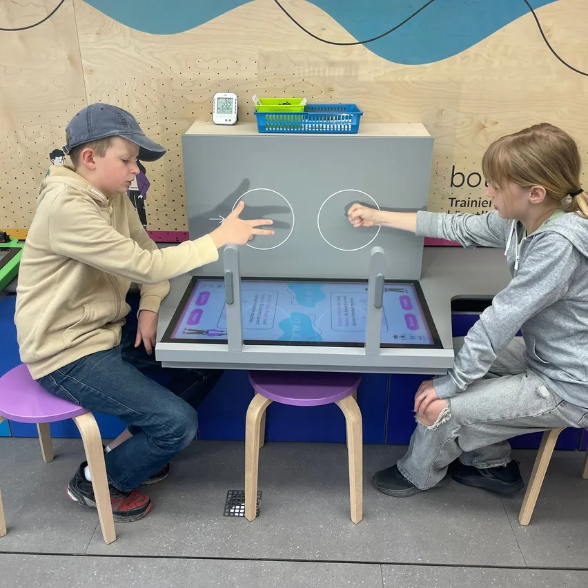 Zwei Kinder interagieren mit einem digitalen Display in einem Workshop. Eines drückt einen Knopf mit dem Finger, während das andere wartet.