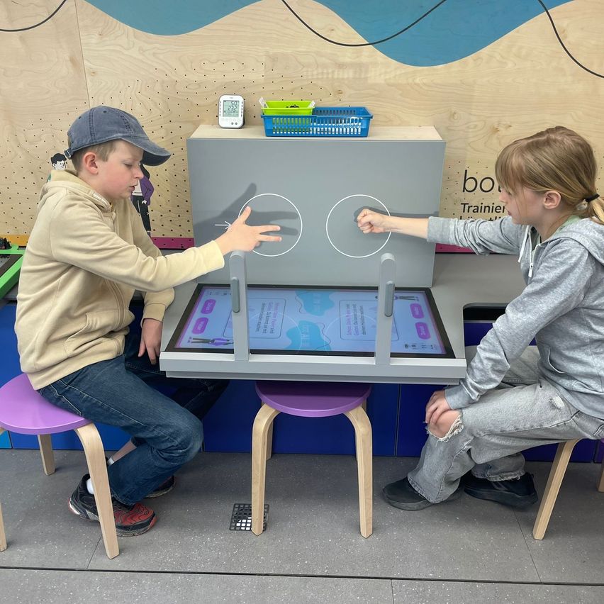 Two children interact with a digital display in a workshop. One presses a button with their finger while the other waits.