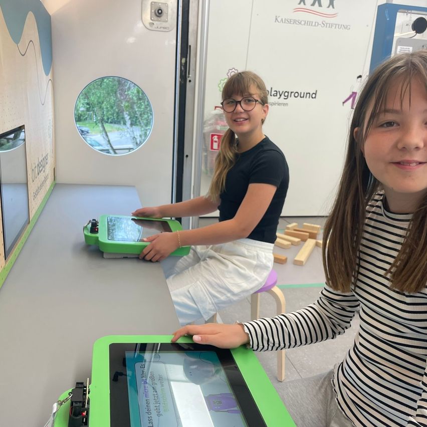 Two girls are sitting at a table with a green tablet in a room with a circular window showing trees outside. One girl is wearing glasses and the other has a striped shirt.