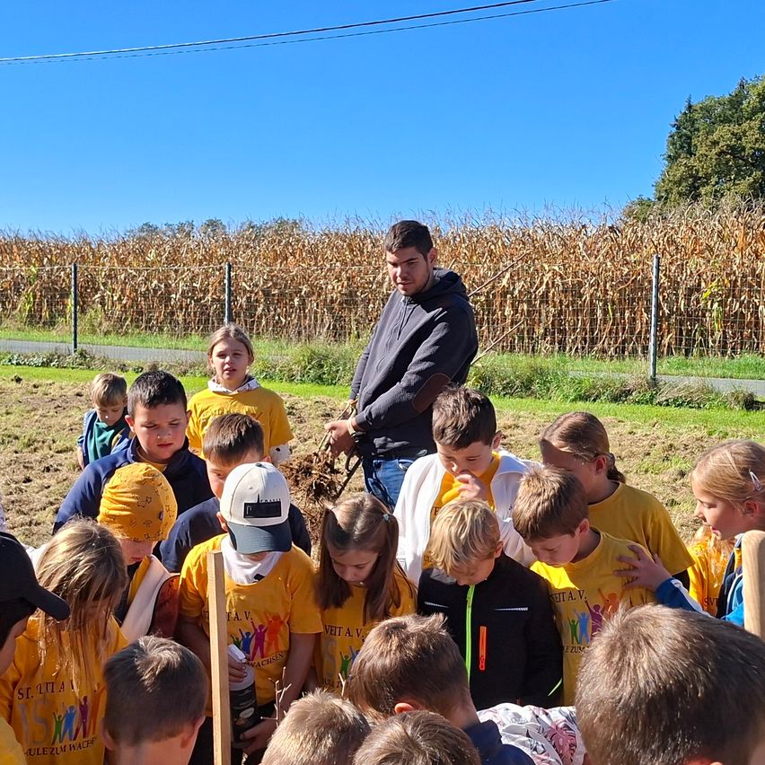 Eine Gruppe von Kindern in gelben T-Shirts ist um einen Mann versammelt, der neben einem Baum in einem Maisfeld steht.