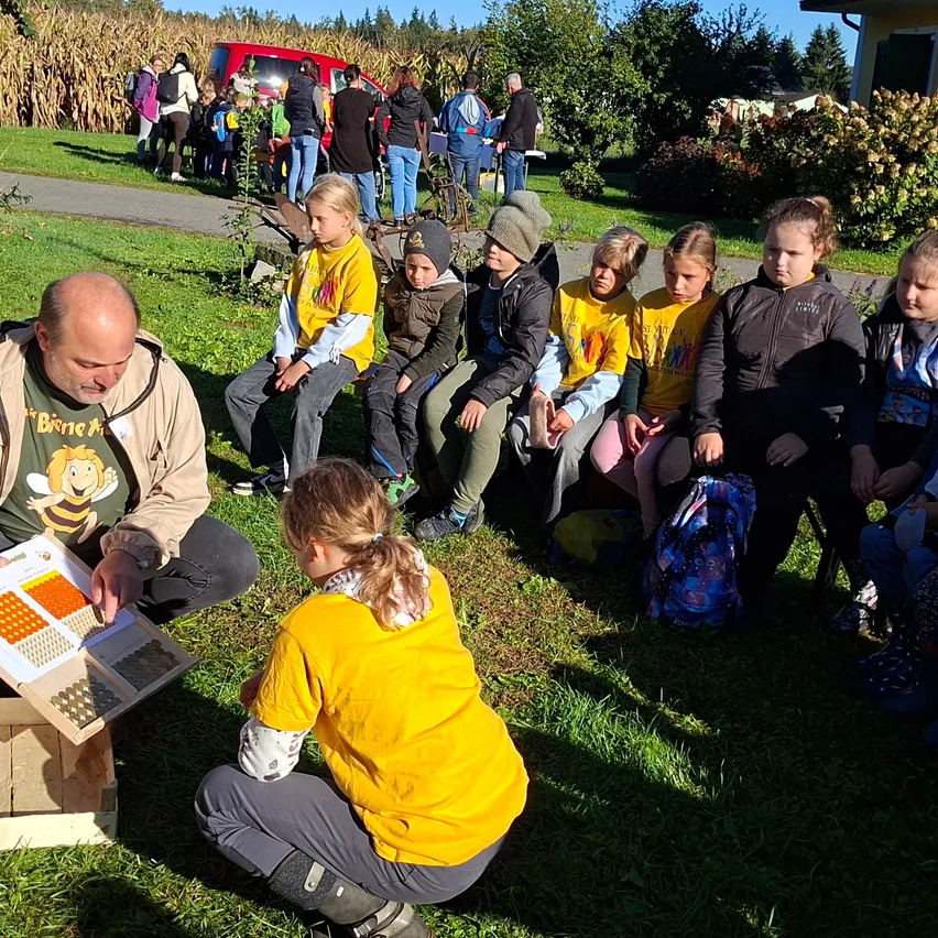 Eine Gruppe von Kindern und Erwachsenen versammelt sich um einen Mann, der auf dem Gras sitzt und ein Buch liest. Die Kinder tragen gelbe Hemden und sitzen auf dem Gras. Es gibt hölzerne Bienenstöcke und andere Gegenstände auf dem Boden.