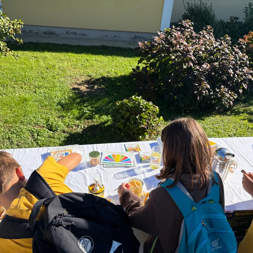 Eine Gruppe von Kindern sitzt draußen an einem Tisch. Sie tragen Rucksäcke und scheinen an einer Aktivität beteiligt zu sein.
