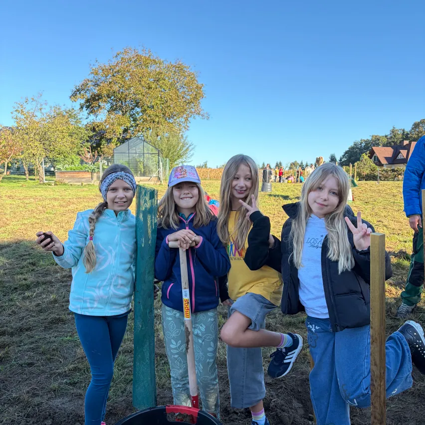 Vier junge Mädchen stehen auf einem Feld und lächeln für ein Foto. Sie halten Schaufeln und stehen neben einem Eimer und einem Holzpfahl.