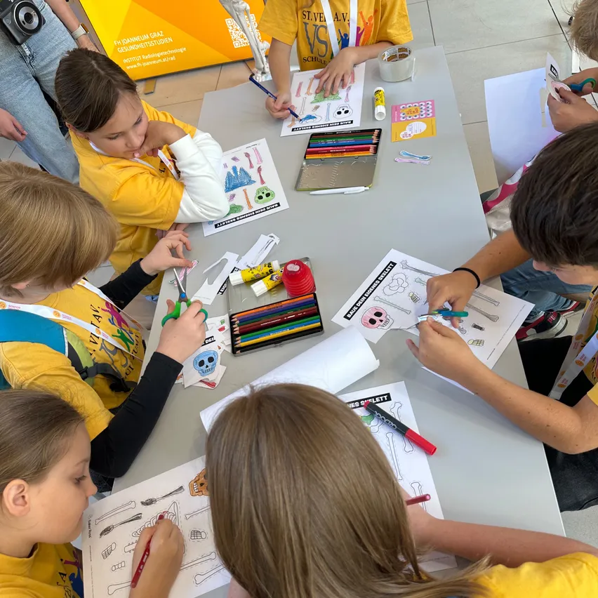 Eine Gruppe von Kindern in gelben T-Shirts sitzt um einen Tisch herum und zeichnet mit Markern und Scheren auf Papier.