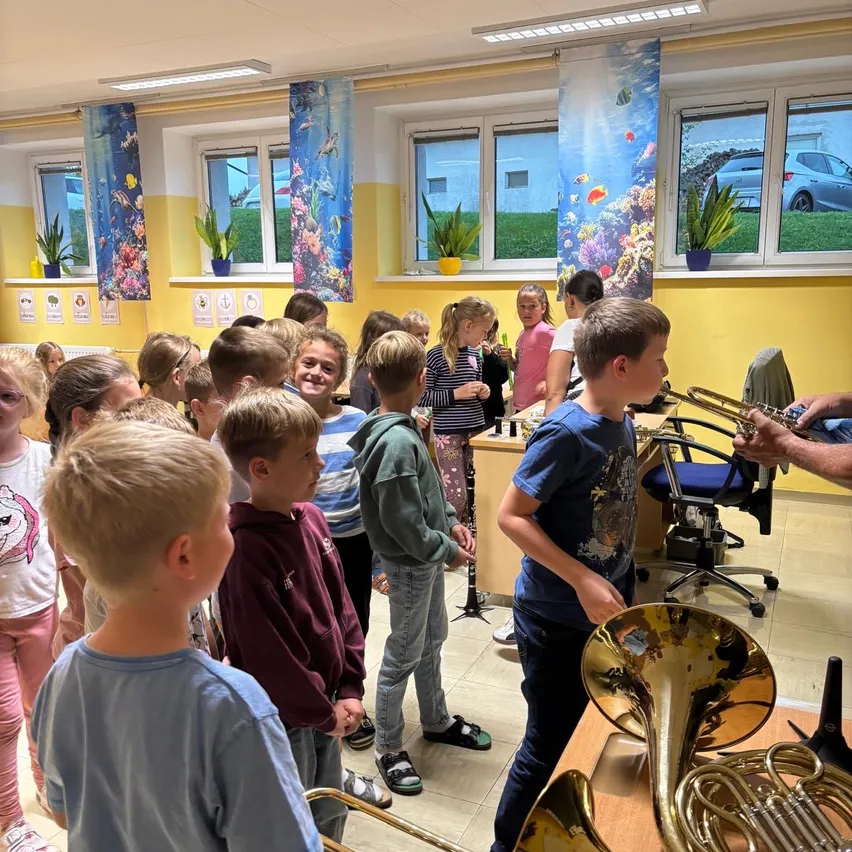 Eine Gruppe von Kindern versammelt sich in einem Klassenzimmer und beobachtet eine Person, die eine Posaune spielt.