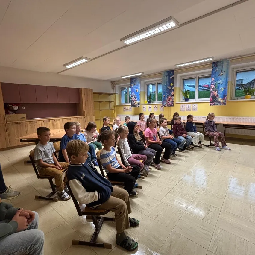 Ein Klassenzimmer mit Kindern, die in Reihen auf Stühlen sitzen und auf eine Vorderwand mit Fenstern und einer Tafel schauen.