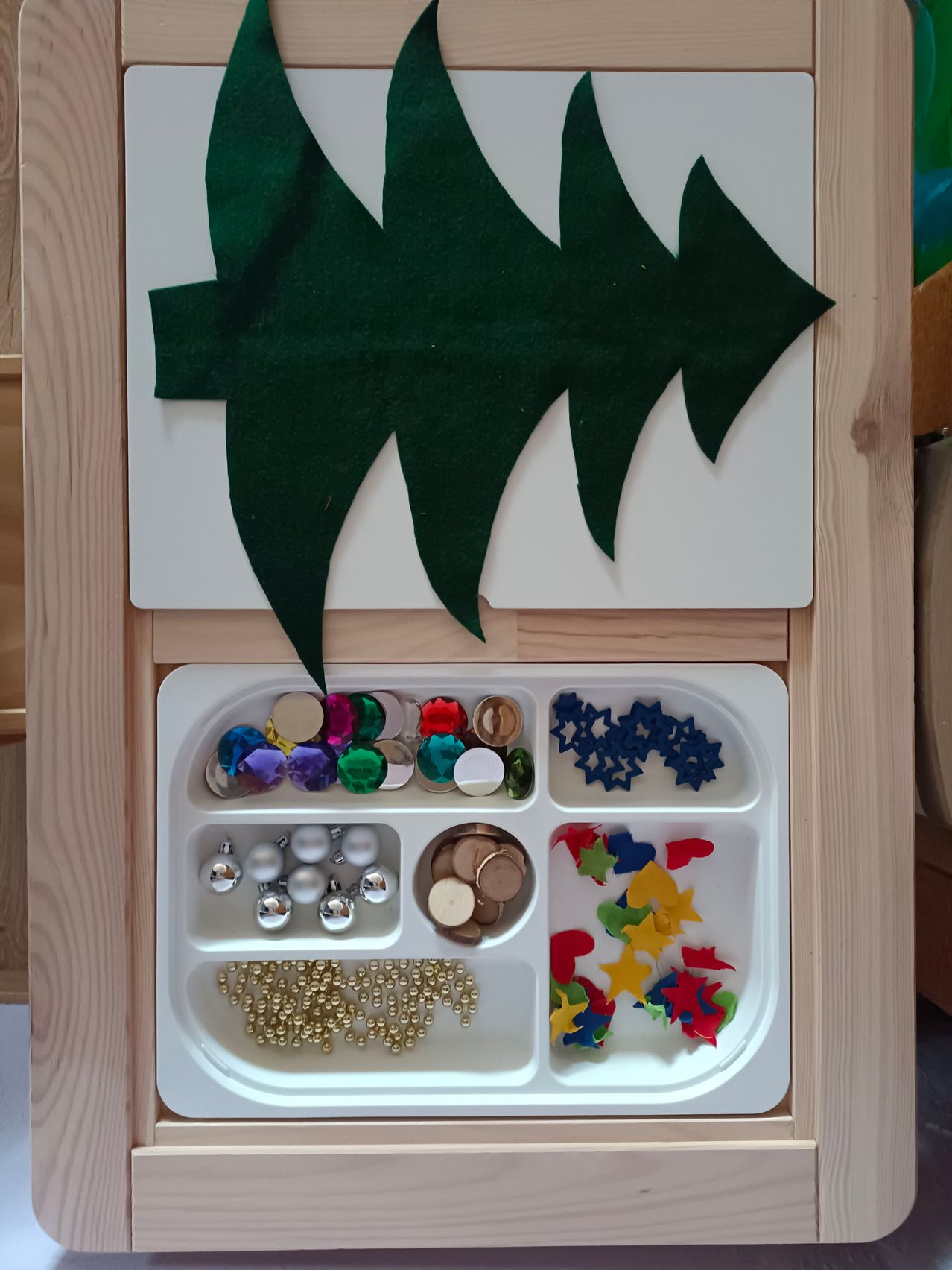 Ein Holztablett mit einem Weihnachtsbaum aus grünem Filz obenauf. Darunter enthält das Tablett verschiedene Dekorationsartikel, darunter bunte Perlen, Sterne und Ornamente.