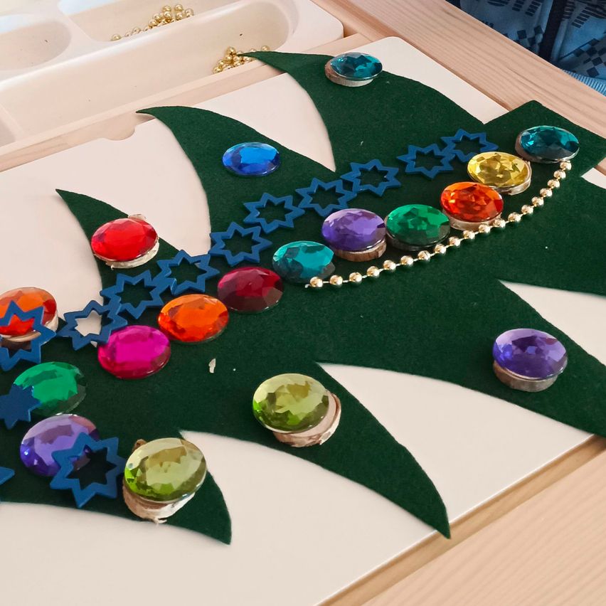 Eine handgefertigte Weihnachtsbaumdekoration mit einer grünen Filzbasis und bunten Edelsteinen. Der Baum hat eine Perlenkette mit einem Sternenmuster und verschiedenen farbigen Edelsteinen.