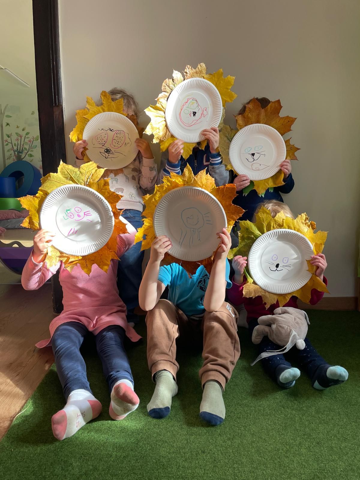 Several children are sitting on the floor, holding paper plates with drawings on them, likely for a photo. They are all wearing socks and some have leaves on their plates.