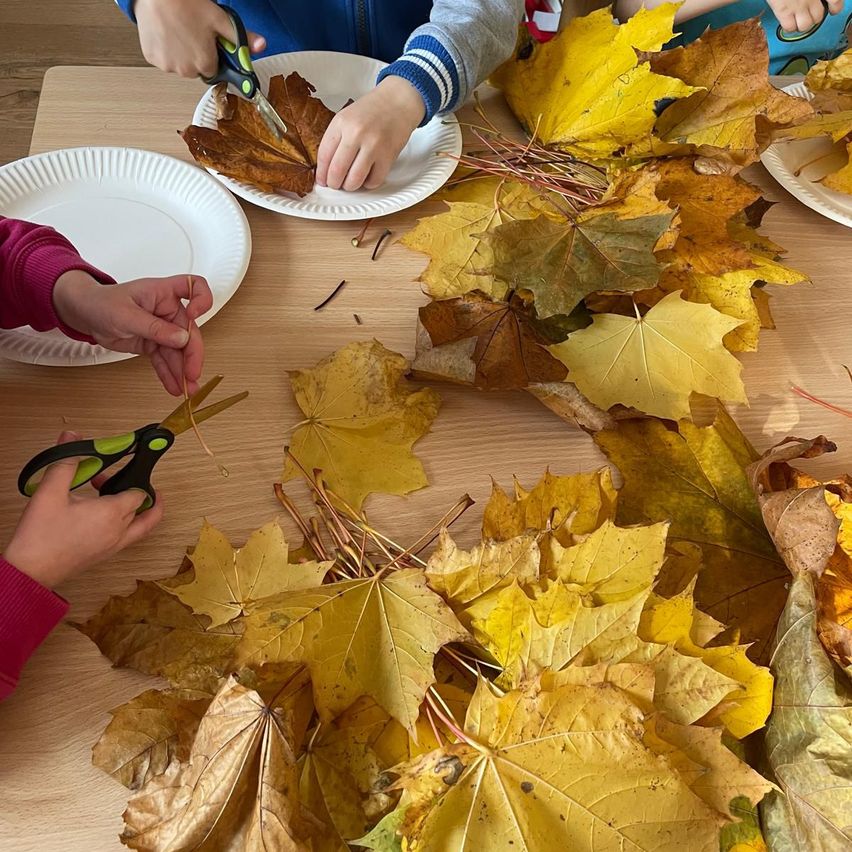 Children are using scissors to cut leaves on a table, with paper plates nearby. The leaves are mostly yellow and brown.