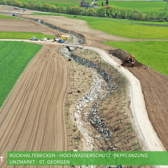 Luftaufnahme einer Baustelle in einem ländlichen Gebiet mit Fahrzeugen, Maschinen und einer Brücke. Grüne und braune Felder sind sichtbar.