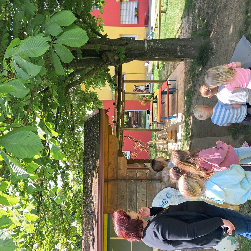 Eine Gruppe von Kindern steht in einem Spielplatz in der Nähe eines Baumes. In der Nähe befindet sich ein Gebäude mit roter Wand, das eine Bank und einen Tisch hat.