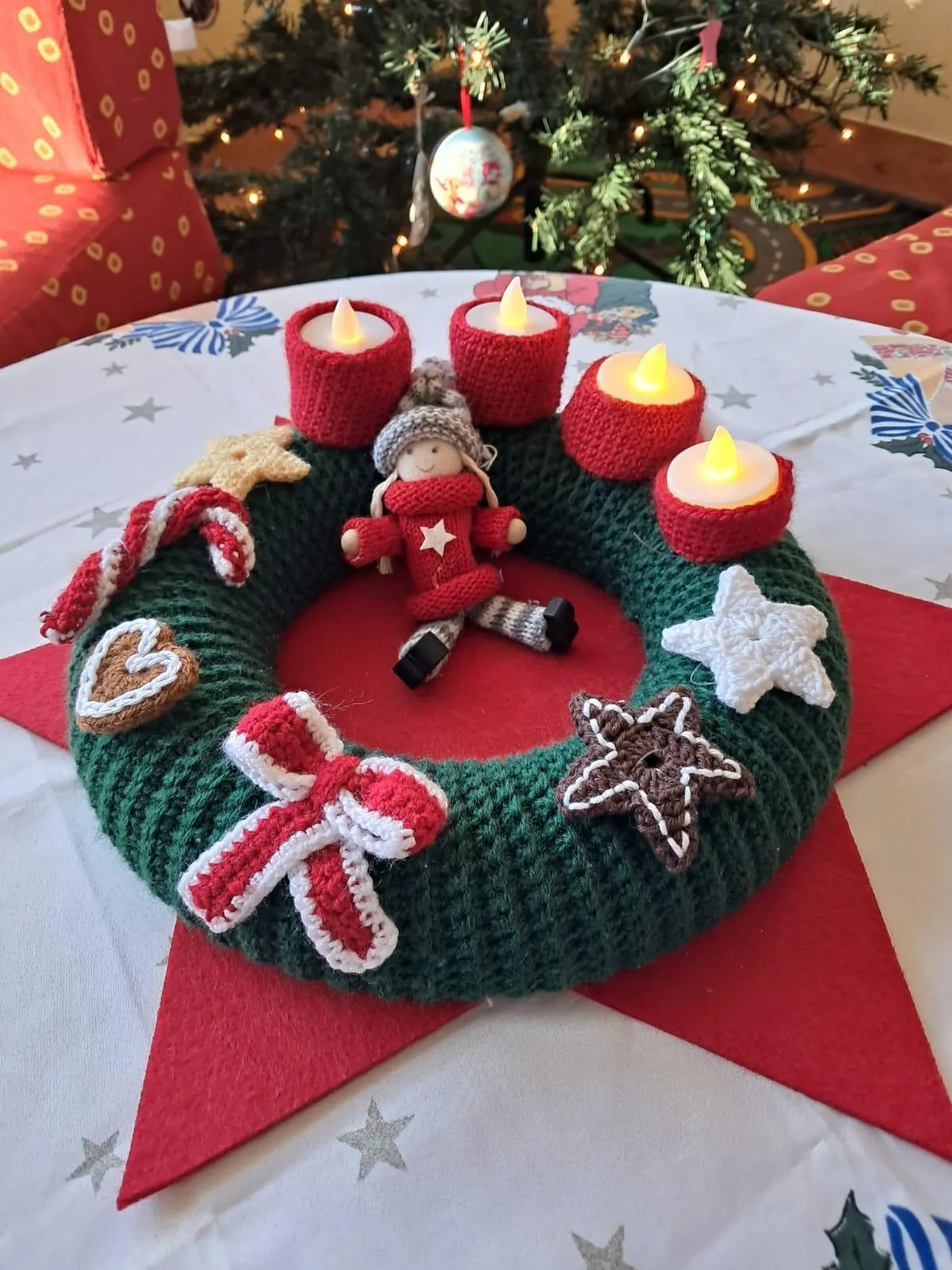 Ein gehäkeltes Adventskranz mit Weihnachtsdekorationen, darunter eine Puppe, Sterne, Kekse und vier brennende Kerzen. Der Kranz steht auf einem Tisch mit einem Weihnachtsbaum im Hintergrund.