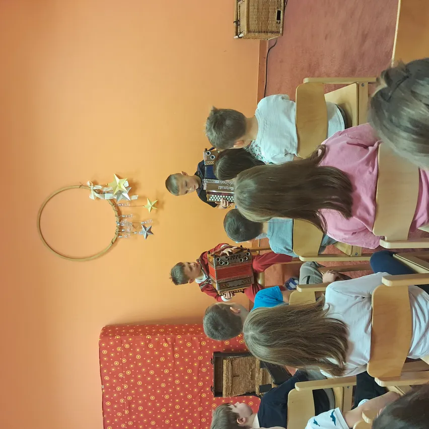 Kinder sitzen in Stühlen in einem Raum, ein Junge spielt Akkordeon. Die Wand hinter ihnen ist orange gestrichen mit einem dekorativen Reifen und Sternen.