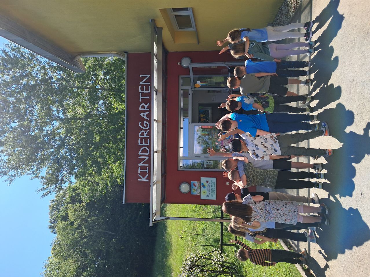 A group of children are standing in a line outside a kindergarten building, with a clear blue sky above and a tree in the background.