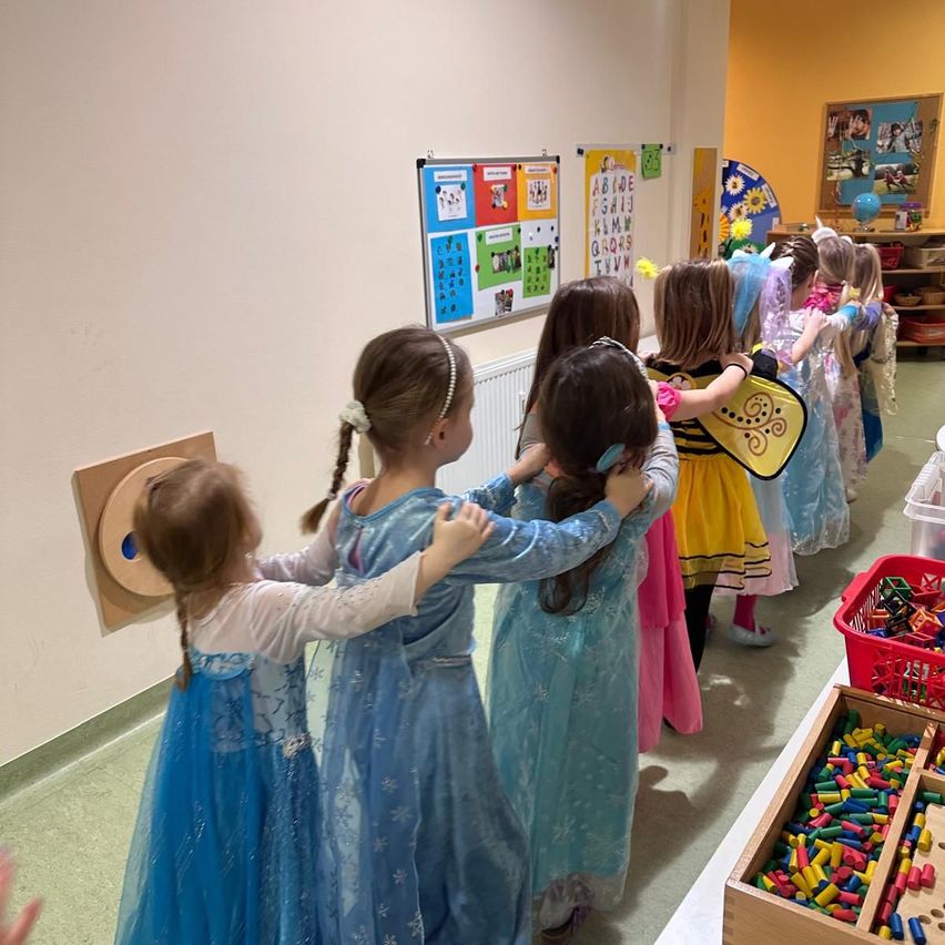 A group of young girls dressed in various costumes, including a bee, stand in a circle, possibly rehearsing for a play in a room with toys and posters.