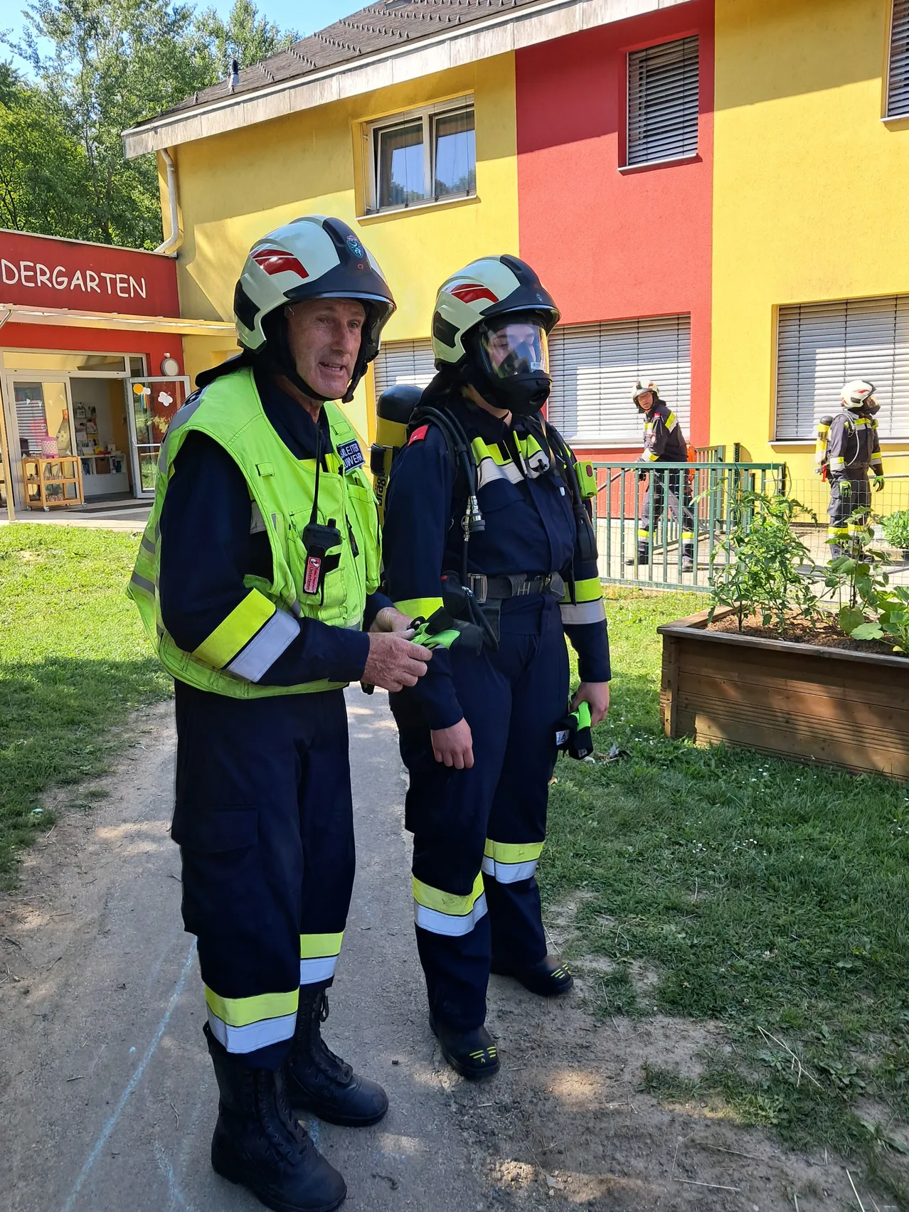 Zwei Feuerwehrleute in voller Ausrüstung stehen vor einem Gebäude, das mit 'Kindergarten' beschriftet ist. Sie scheinen zu sprechen. Im Hintergrund sind weitere Feuerwehrleute zu sehen.