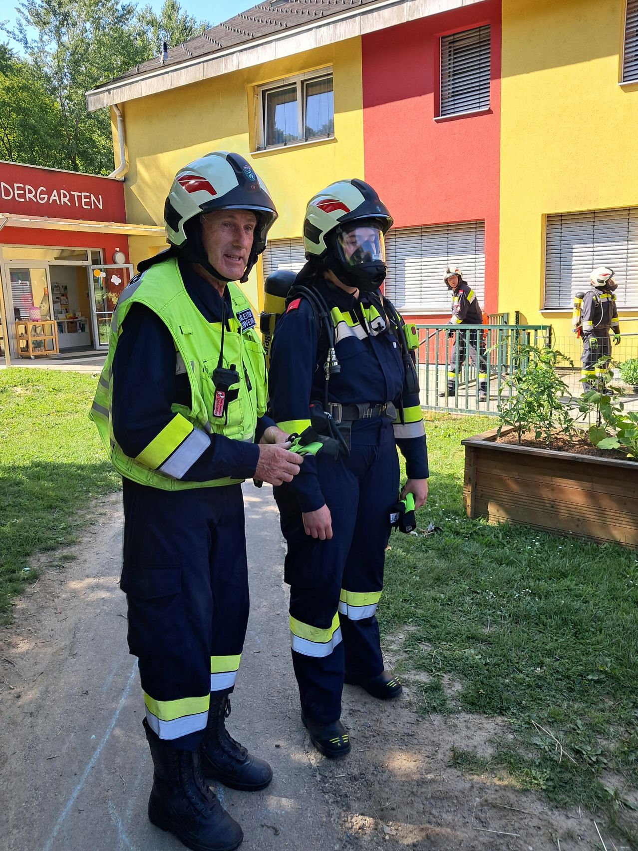 Zwei Feuerwehrleute in voller Ausrüstung stehen vor einem Gebäude, das mit 'Kindergarten' beschriftet ist. Sie scheinen zu sprechen. Im Hintergrund sind weitere Feuerwehrleute zu sehen.
