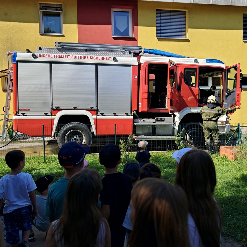 Eine Gruppe von Kindern versammelt sich um einen roten Feuerwehrwagen. Ein Feuerwehrmann erklärt ihnen etwas, während er neben dem Wagen steht. Dahinter befindet sich ein gelbes Gebäude mit Fenstern.