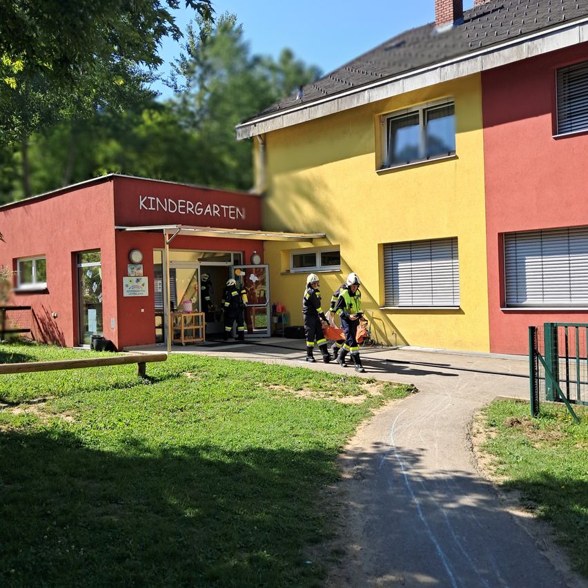 Drei Feuerwehrleute in Uniform evakuieren einen Kindergarten. Sie tragen einen orangefarbenen Beutel und gehen den Weg hinunter.