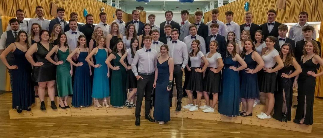 Eine Gruppe formell gekleideter Personen steht auf einem Holzboden, möglicherweise für ein Foto. Sie sind in verschiedenen Farben und Stilen gekleidet.