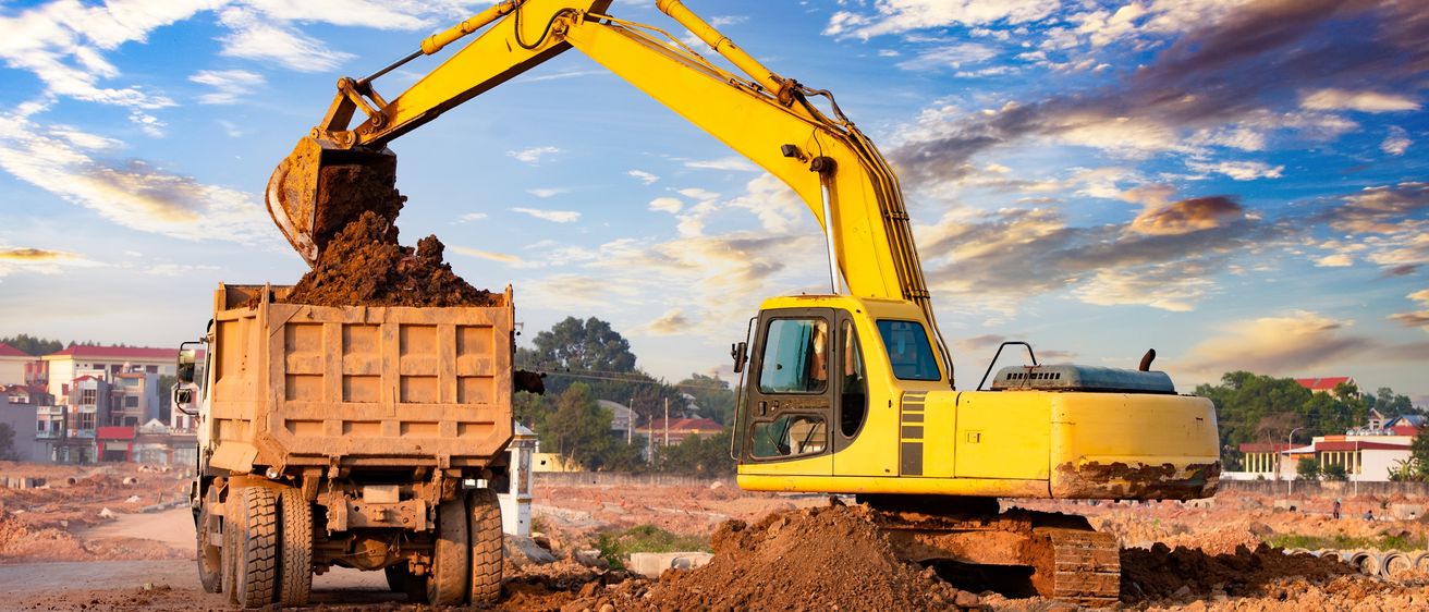 Ein gelber Bagger kippt Erde in einen Kipplaster auf einer Baustelle unter blauem Himmel mit Wolken aus.