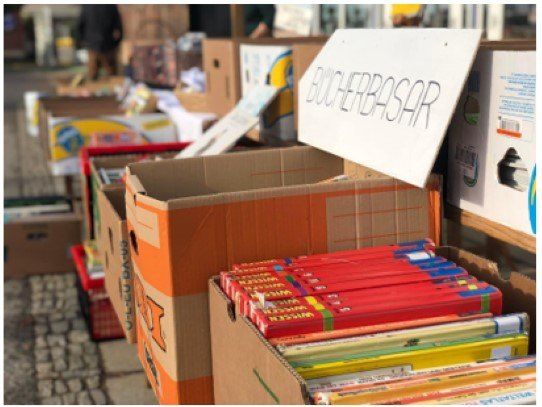 Stapel von Kartons mit Büchern, ein Schild oben drauf sagt 'BUCHERBASAR'. Die Kartons sind offen, und einige Bücher sind aufeinandergestapelt. Ein roter Kasten ist links.