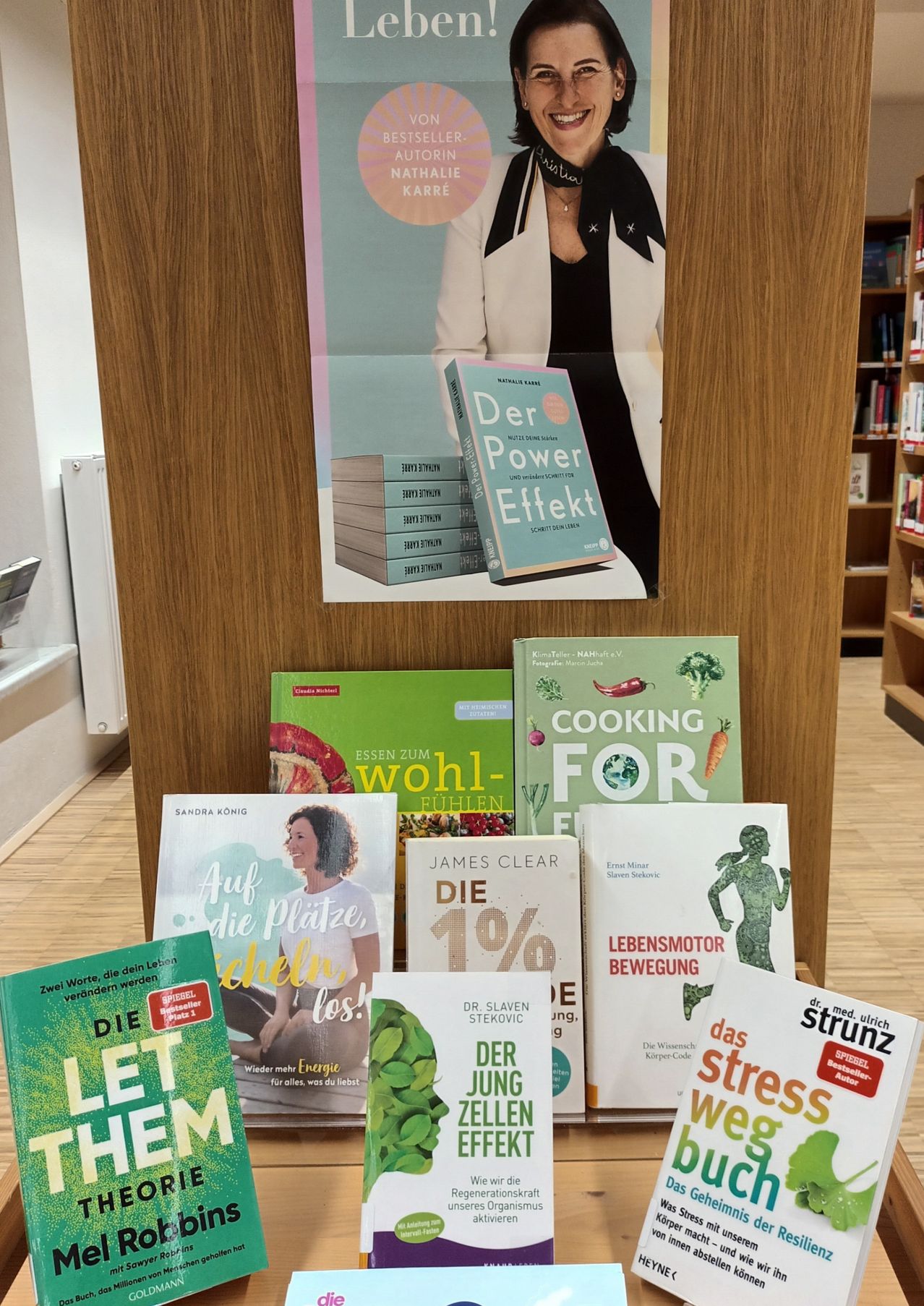 Eine Buchausstellung mit verschiedenen Titeln, darunter 'Der Power Effekt', 'Auf die Platze, chelen los!' und 'Lebensmotor Bewegung', mit einem Poster einer Frau in einer weißen Jacke.