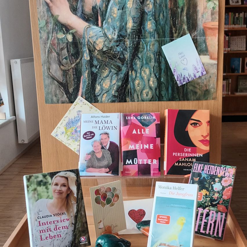 Ein hölzerner Tisch zeigt mehrere Bücher, darunter 'Meine Mutter' und 'Meine Mama Die Lowin'. Im Hintergrund befinden sich ein großes Gemälde und ein Regal mit Büchern. Eine Notiz ist an das Gemälde geheftet.