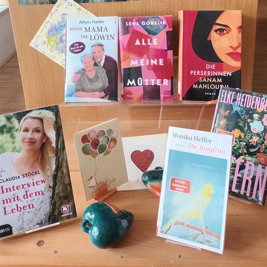 Verschiedene Bücher sind auf einer hölzernen Oberfläche ausgestellt. Titel beinhalten 'Meine Mama die Lowin' und 'Die Personinnen Sanam Mahloudi'. Ein grüner apfelförmiger Gegenstand ist in der Mitte.