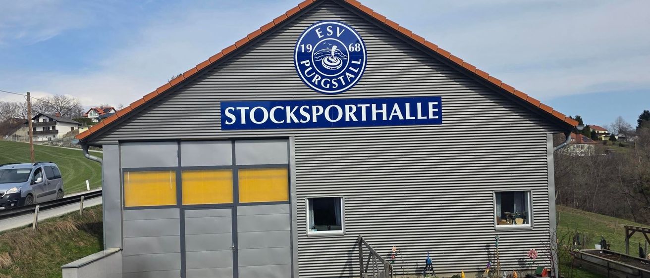 Ein graues Gebäude mit einem blauen Schild, das 'Stocksporthalle' und ein Logo mit 'ESV Purgstall 68' zeigt. Es hat eine Garagentür und Fenster.