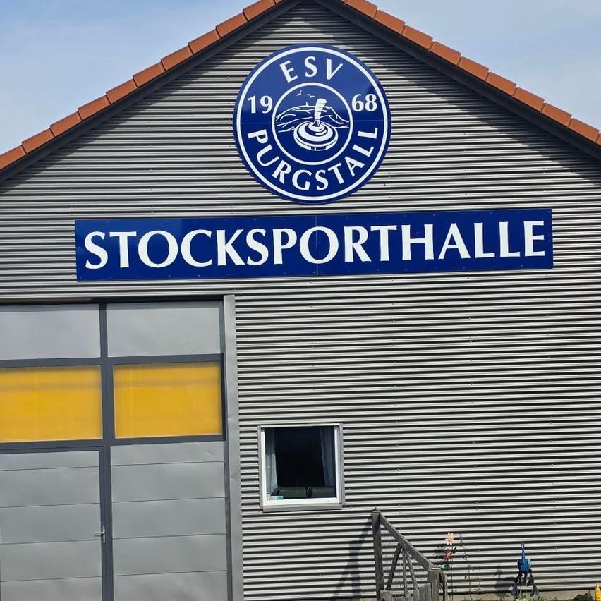 Ein graues Gebäude mit einem blauen Schild, das 'Stocksporthalle' und ein Logo mit 'ESV Purgstall 68' zeigt. Es hat eine Garagentür und Fenster.