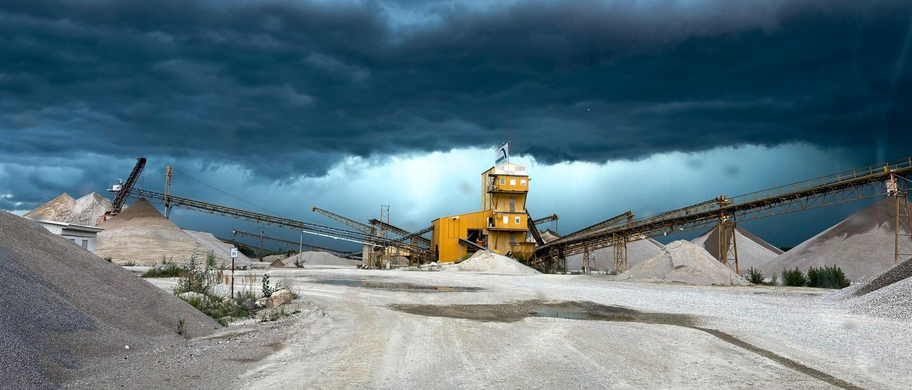 Ein stürmischer Himmel überlagert einen industriellen Steinbruch. Eine große gelbe Maschinenstruktur steht prominent, mit Förderbändern, die Materialien transportieren. Der Boden ist mit Kies und Erde übersät.
