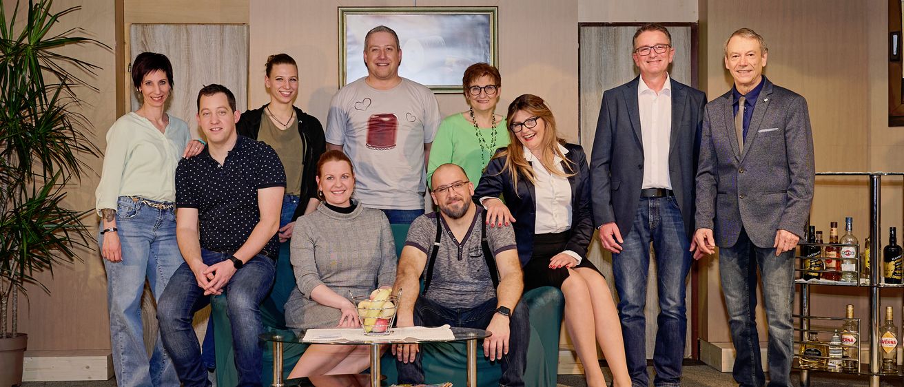 Eine Gruppe von Männern und Frauen, die lässig gekleidet sind, posiert für ein Foto auf einer Bühne mit Hintergrund. Ein Tisch mit Obst und Zeitschriften steht davor.