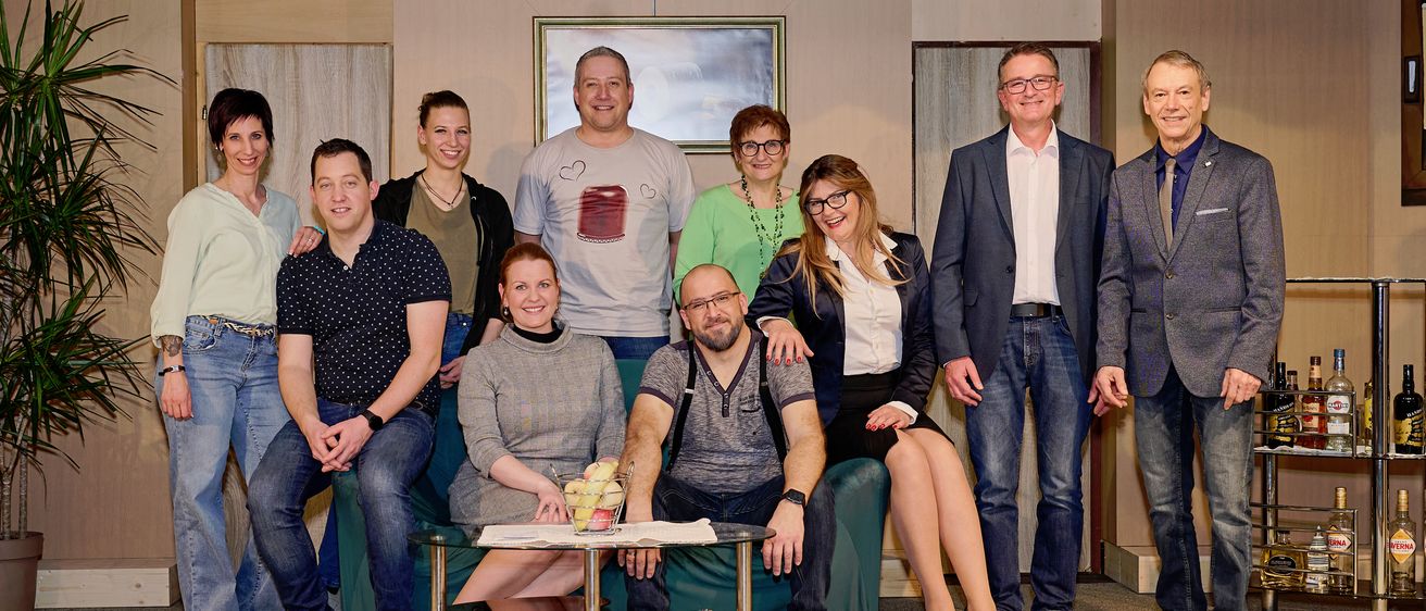 Eine Gruppe von Männern und Frauen, die lässig gekleidet sind, posiert für ein Foto auf einer Bühne mit Hintergrund. Ein Tisch mit Obst und Zeitschriften steht davor.