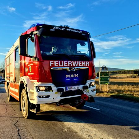 Ein roter MAN-Feuerwehrwagen fährt auf einer Straße unter blauem Himmel mit grünen Schildern an der Seite.