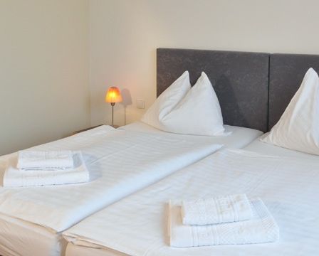 Zwei ordentlich gemachte Betten mit weißen Laken, Kissen und Handtüchern in einem sauberen, weißen Schlafzimmer mit grauen Kopfteilen. Eine kleine Lampe steht auf dem Nachttisch.