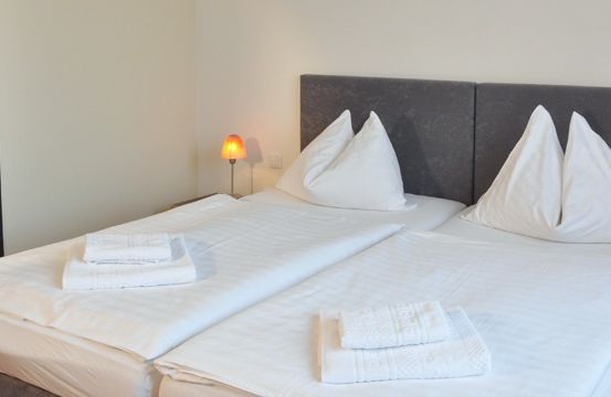 Zwei ordentlich gemachte Betten mit weißen Laken, Kissen und Handtüchern in einem sauberen, weißen Schlafzimmer mit grauen Kopfteilen. Eine kleine Lampe steht auf dem Nachttisch.
