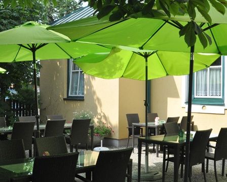 Ein Restaurantbereich mit grünen Sonnenschirmen und Stühlen um Tische herum, mit einem gelben Gebäude im Hintergrund.