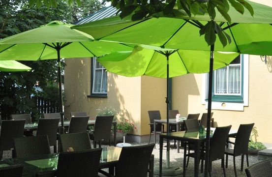Ein Restaurantbereich mit grünen Sonnenschirmen und Stühlen um Tische herum, mit einem gelben Gebäude im Hintergrund.