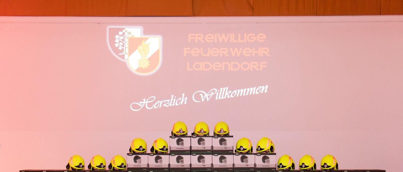 Eine Ausstellung von gelben Helmen, die in Reihen auf einer Bühne mit einem rosa Hintergrund angeordnet sind, einschließlich Logos und Sponsoren für Freiwillige Feuerwehr Ladendorf.