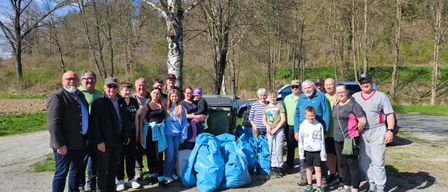 Eine Gruppe von Menschen in Freizeitkleidung posiert für ein Foto, einige halten blaue Taschen, in einem Waldgebiet mit Bäumen, Mülleimern und einem Fahrzeug im Hintergrund.